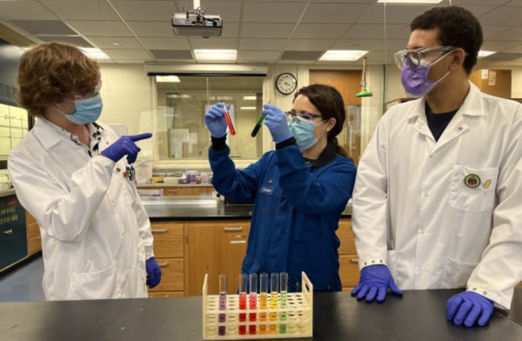 3 students in a lab with a rack of test tubes on a table. One student is holding up 2 test tubes, while the other two students look on.